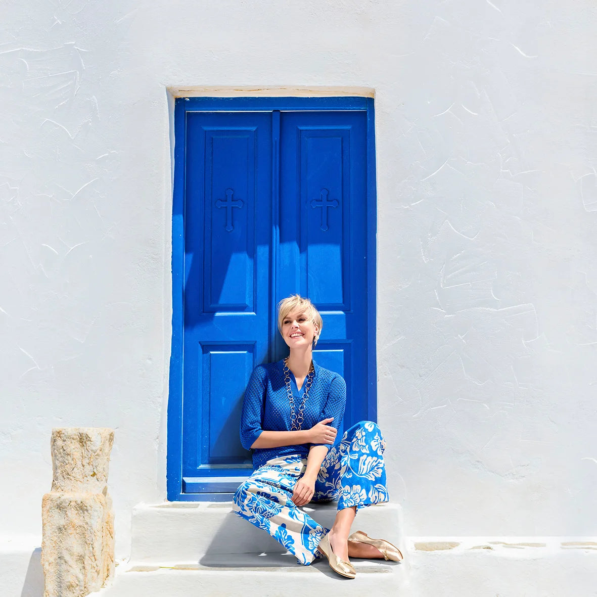 A model sitting in front of a blue door, wearing blue trousers and a blue polo pull with crochet