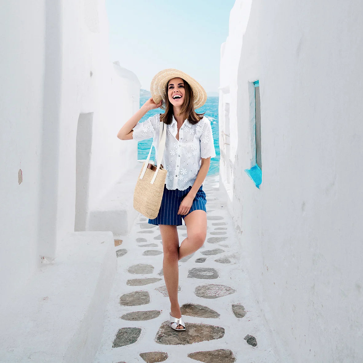A model wearing navy striped shorts and a white broderie Anglaise blouse while standing in a Greek alley in front of the sea